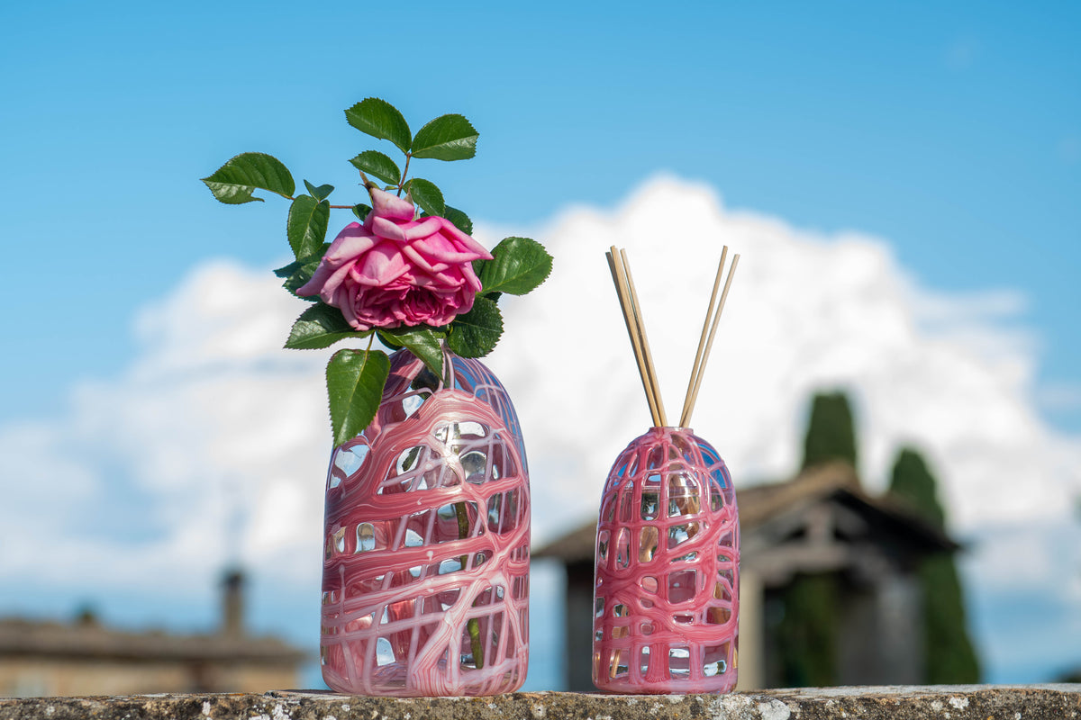 A pink and clear woven decorative vase holding a pink rose with green leaves, standing beside a matching pink and clear reed diffuser with wooden sticks, set outdoors against a blue sky and blurred building background.