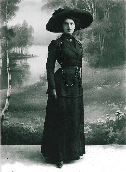 Archival picture depicting Woman in a dark formal dress and hat standing in front of a scenic backdrop